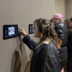 Zwischen Seidenstraße und Gegenwartskunst 4 Mehrere junge Erwachsene stehen in einer Ausstellung an einer Wand mit montierten Tablets. Eine Person im Vordergrund mit Kopfhörern und dunkler Lederjacke tipp