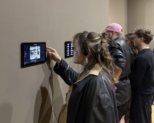 Besucherinnen und Besucher interagieren mit Tablets in einer Medienausstellung Mehrere junge Erwachsene stehen in einer Ausstellung an einer Wand mit montierten Tablets. Eine Person im Vordergrund mit Kopfhörern und dunkler Lederjacke tipp