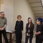 Zwischen Seidenstraße und Gegenwartskunst 6 Vier Personen stehen in einem Treppenhaus und unterhalten sich. Links steht ein Mann mit Brille und grauem Haar in einem olivgrünen Hemd, daneben zwei Frauen in