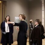 Zwischen Seidenstraße und Gegenwartskunst 10 Eine Frau in schwarzem Kleid führt eine kleine Gruppe von Besucherinnen durch einen Ausstellungsraum und zeigt mit ausgestrecktem Arm auf ein Objekt außerhalb d
