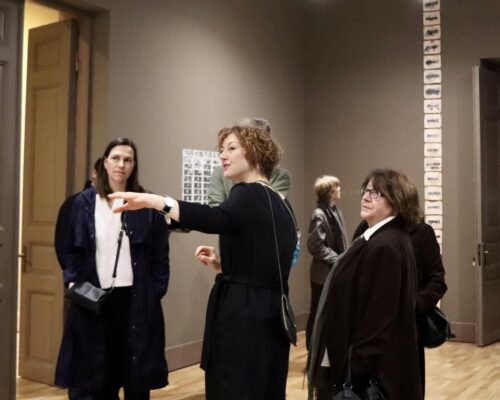 Führung durch eine Kunstausstellung Eine Frau in schwarzem Kleid führt eine kleine Gruppe von Besucherinnen durch einen Ausstellungsraum und zeigt mit ausgestrecktem Arm auf ein Objekt außerhalb d