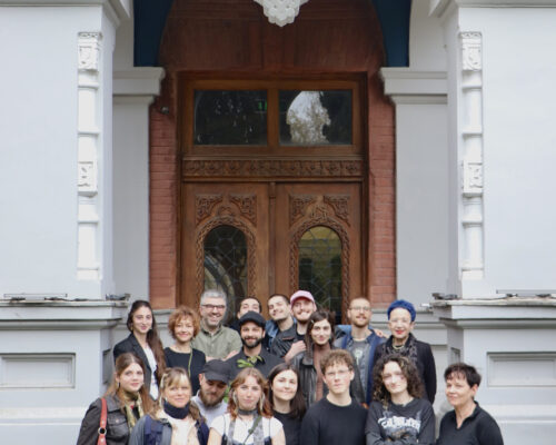 Gruppe vor historischem Gebäude mit Holztor und Blumenwiese Gruppenfoto von etwa zwanzig Menschen unterschiedlichen Alters vor einem großen, dekorativen Holztor eines historischen Gebäudes. Die Gruppe steht eng beisammen