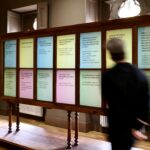 Zwischen Seidenstraße und Gegenwartskunst 7 Person in schwarzer Kleidung geht an einer großen hölzernen Vitrine in einem Ausstellungsraum vorbei. In der Vitrine sind zahlreiche farbige Texttafeln mit phil