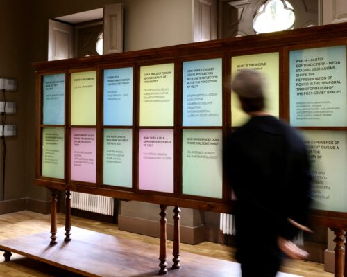 Besucher vor Textinstallation in einem Museum Person in schwarzer Kleidung geht an einer großen hölzernen Vitrine in einem Ausstellungsraum vorbei. In der Vitrine sind zahlreiche farbige Texttafeln mit phil