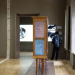 Zwischen Seidenstraße und Gegenwartskunst 8 Innenraum einer modernen Kunstausstellung mit hellem Holzboden. In der Mitte steht ein hohes Holzobjekt mit drei farbigen Texttafeln in Englisch und einer weite