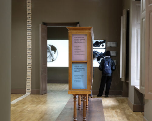 Zeitgenössische Kunstausstellung mit installativen Text- und Videoprojektionen Innenraum einer modernen Kunstausstellung mit hellem Holzboden. In der Mitte steht ein hohes Holzobjekt mit drei farbigen Texttafeln in Englisch und einer weite
