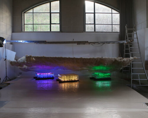 Schwebende Wolkenskulptur über farbig beleuchteten Lichtkästen im Atelier Innenraum eines Ateliers mit hohen, bogenförmigen Fenstern. In der Mitte hängt eine lange, wolkenartige Skulptur aus Watte oder Stoff knapp über dem Boden. Daru