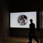 Zwischen Seidenstraße und Gegenwartskunst 11 Abgedunkelter Ausstellungsraum mit einer großen, leuchtenden Projektionsfläche an der Wand, auf der ein rundes Schwarz-Weiß-Bild einer Person zu sehen ist, die