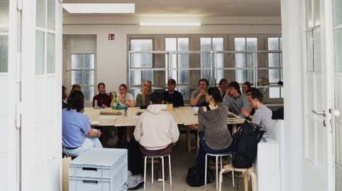 Eine Gruppe von etwa fünfzehn Personen sitzt um einen großen rechteckigen Holztisch in einem hellen Seminarraum. Alle blicken nach vorn, einige machen sich Noti