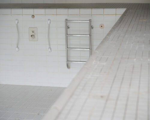 Leeres, weiß gefliestes Schwimmbecken mit Metallleiter an der Seitenwand und sichtbarem Beckenrand im Vordergrund.