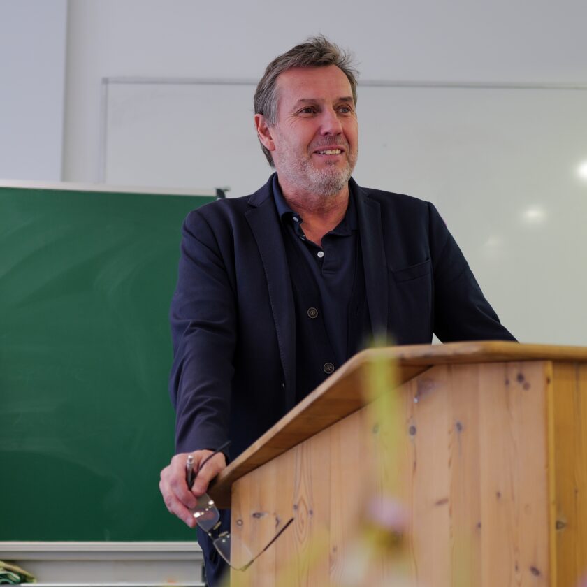 Mittelalter Mann in Sakko und Polohemd steht lächelnd hinter einem hölzernen Pult in einem Klassenraum, vor einer grünen Tafel und einer weißen Wandtafel, und h