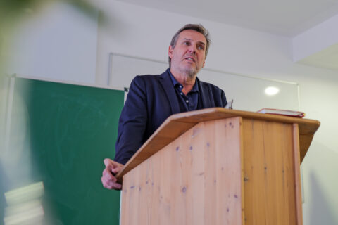 Ein Mann mittleren Alters steht in einem Seminarraum hinter einem hölzernen Rednerpult und spricht, im Hintergrund sind eine grüne Tafel und ein Whiteboard zu s