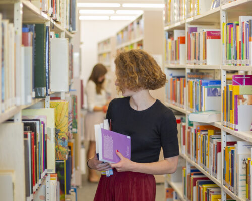Studierende sucht nach Literatur in einer Bibliothek Junge Frau mit lockigen Haaren steht zwischen hohen Bücherregalen in einer Bibliothek und hält mehrere Bücher im Arm, während sie zur Seite blickt
