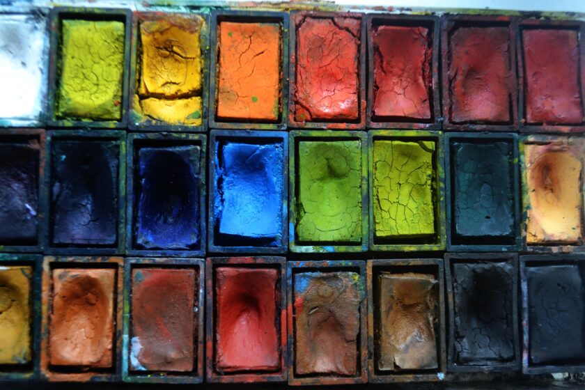 Nahaufnahme eines alten Malkastens mit vielen rechteckigen Farbnäpfen in kräftigen Tönen wie Gelb, Orange, Rot, Blau, Grün, Braun und Schwarz. Die Wasserfarben