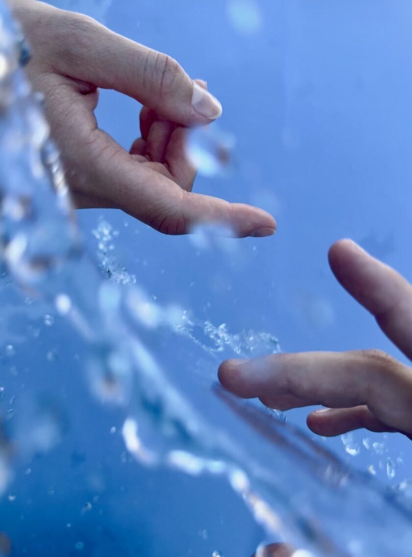Zwei menschliche Hände, die sich vor einem blauen Hintergrund beinahe mit den Fingerspitzen berühren, getrennt durch eine transparente, wellige Oberfläche mit W