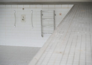 Leeres, weiß gefliestes Schwimmbecken mit Metallleiter an der Wand und Handgriffen, fotografiert aus der Perspektive des Beckenrands.