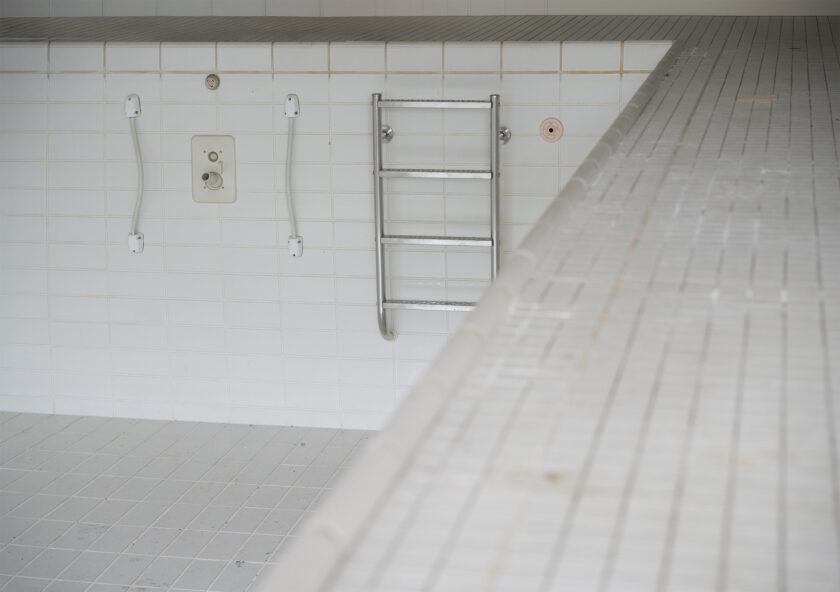 Leeres, weiß gefliestes Schwimmbecken mit Metallleiter an der Wand und Handgriffen, fotografiert aus der Perspektive des Beckenrands.