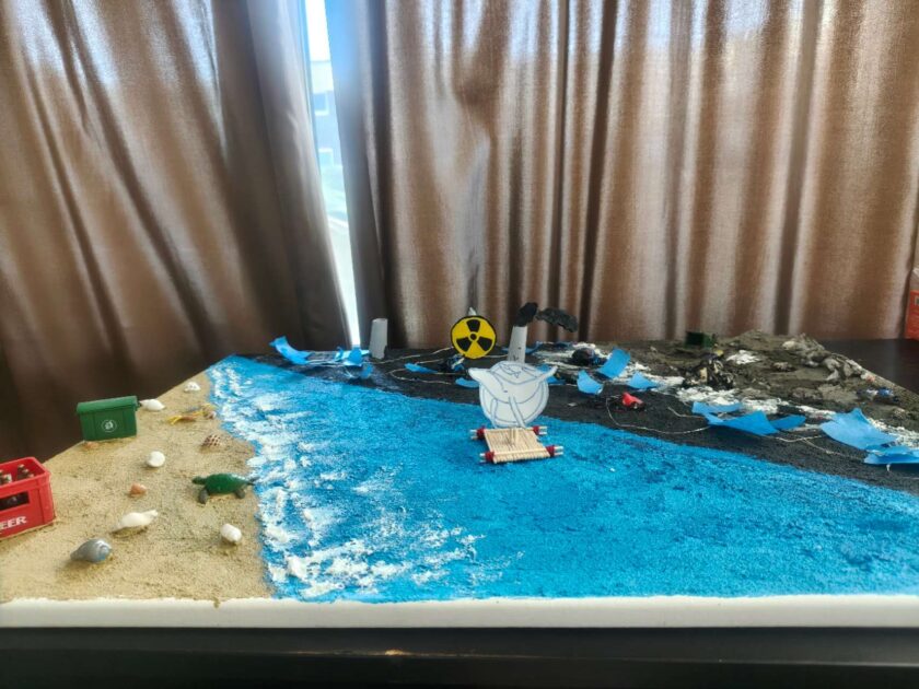 Diorama eines Meeresstrandes mit starkem Kontrast zwischen sauberem und verschmutztem Bereich: Links ein gelber Sandstrand mit Muscheln, einer kleinen Spielzeug