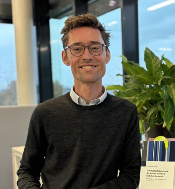 Lächelnder Mann mit Brille und dunklem Pullover steht in einem modernen Büro vor einem großen Fenster, neben ihm eine grüne Zimmerpflanze und ein aufgestelltes