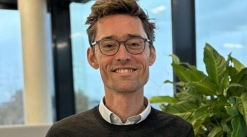 Porträt eines lächelnden Mannes mit kurzer brauner Frisur, Brille und hellblauem Hemd vor neutralem Hintergrund.