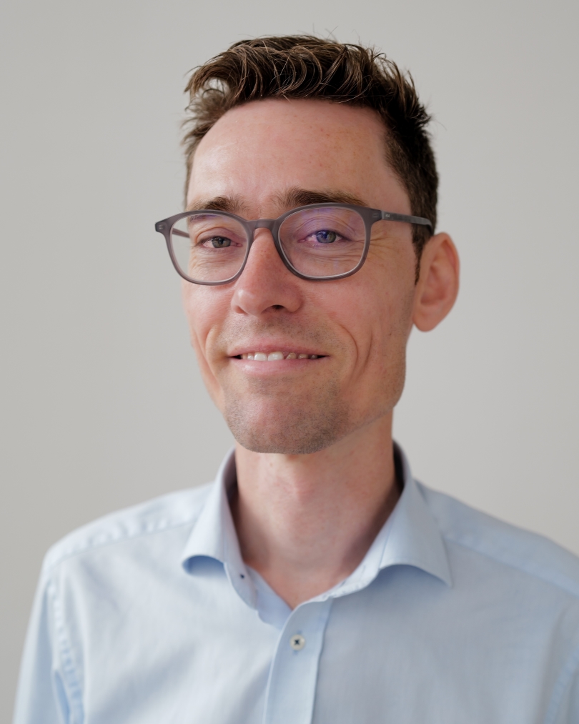 Porträt eines lächelnden Mannes mit kurzer brauner Frisur, Brille und hellblauem Hemd vor neutralem Hintergrund.