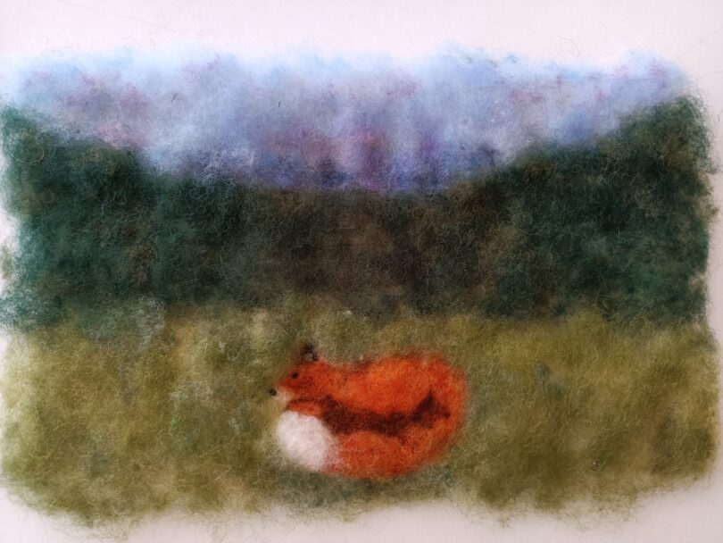 Filzbild mit einer Landschaft: Im Vordergrund liegt ein zusammengerollter orangefarbener Fuchs mit weißer Brust auf einer grünlichen Wiese. Dahinter erstreckt s