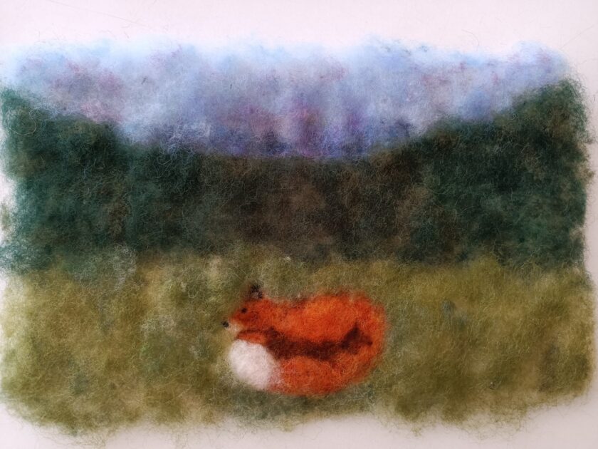 Filzbild mit einer Landschaft: Im Vordergrund liegt ein zusammengerollter orangefarbener Fuchs mit weißer Brust auf einer grünlichen Wiese. Dahinter erstreckt s