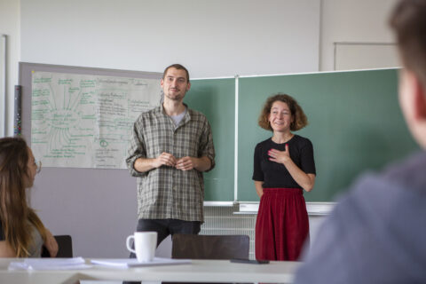Zwei Personen stehen vorne in einem Seminarraum vor einer grünen Tafel und einem Flipchart mit vielen Notizen. Sie sprechen zu einer kleinen Gruppe, die im Vord