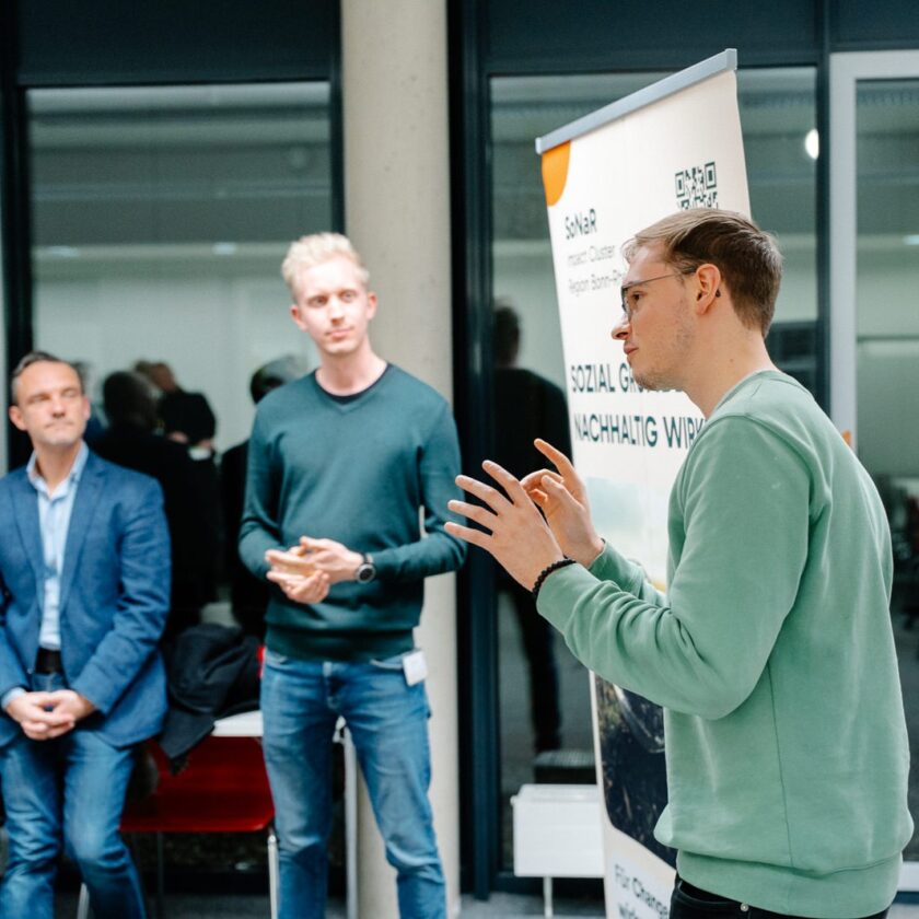 Zwei Männer in einem modernen Innenraum, einer spricht gestikulierend vor einem Roll-up-Banner, der andere hört im Hintergrund zu.