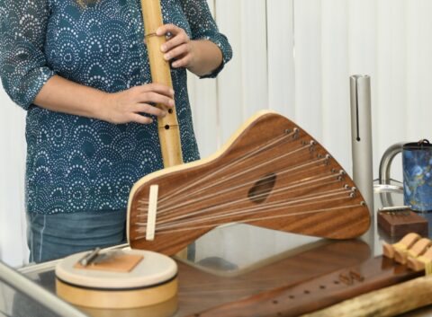 Person in gemusterter blauer Bluse hält eine lange Bambusflöte, vor ihr liegen mehrere Saiteninstrumente aus Holz und ein kleines Trommel-ähnliches Percussionin
