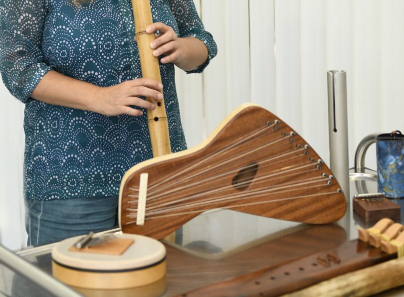 Person in gemusterter blauer Bluse hält eine lange Bambusflöte, vor ihr liegen mehrere Saiteninstrumente aus Holz und ein kleines Trommel-ähnliches Percussionin