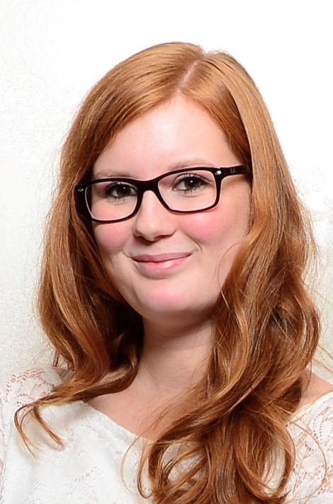 Porträt einer jungen Frau mit langen, welligen, rötlichen Haaren und schwarzer Brille, die leicht lächelnd vor hellem Hintergrund in die Kamera blickt.