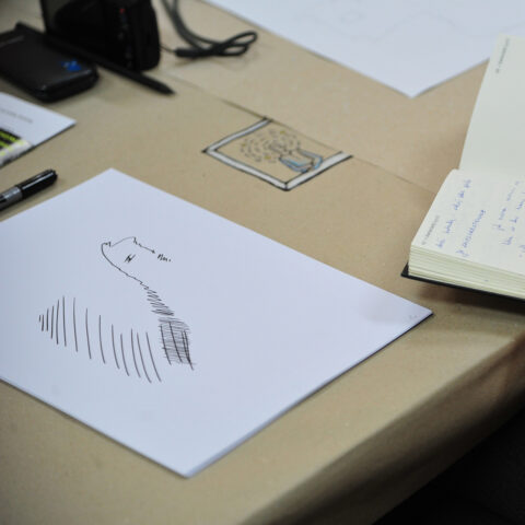 Schreibtisch mit einem Blatt Papier, auf dem einfache schwarze Linien und Skizzen gezeichnet sind, daneben ein offenes Notizbuch mit handschriftlichen Notizen s