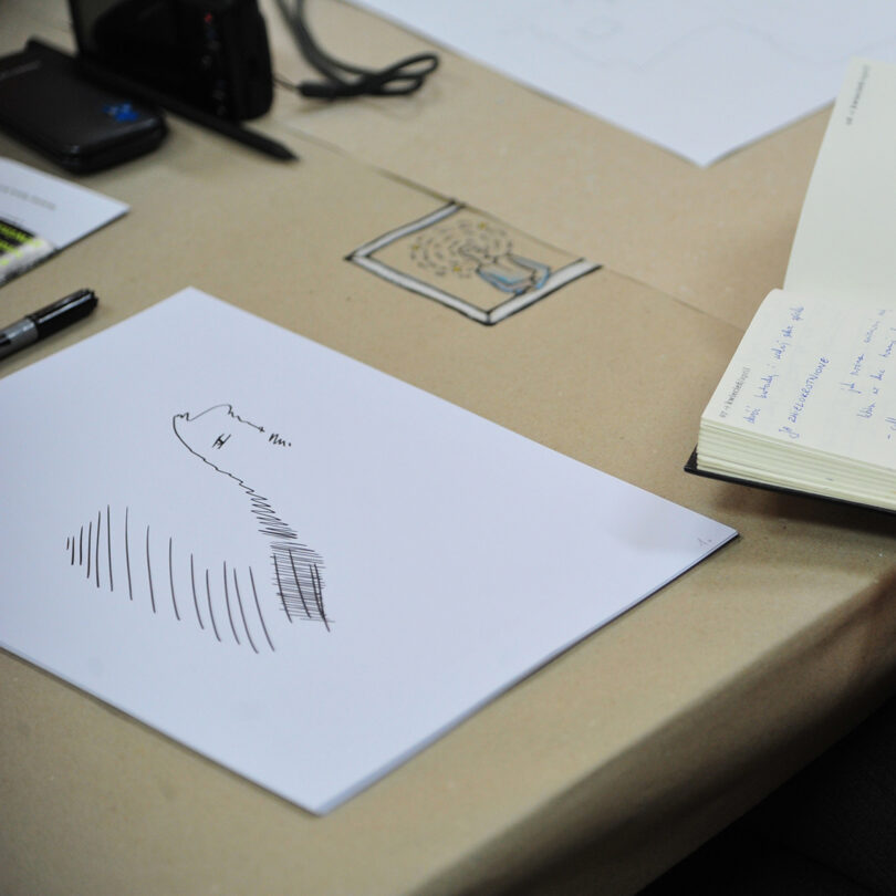 Schreibtisch mit einem Blatt Papier, auf dem einfache schwarze Linien und Skizzen gezeichnet sind, daneben ein offenes Notizbuch mit handschriftlichen Notizen s