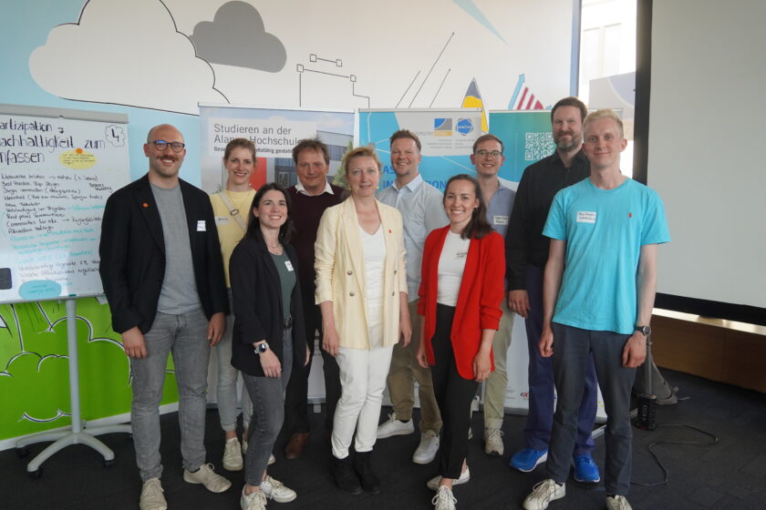 Gruppenfoto von zehn lächelnden Personen in einem modernen Seminarraum vor bunten Roll‑Ups und einem Whiteboard mit Notizen zum Thema Nachhaltigkeit. Die Teilne