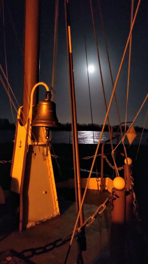 Nächtliche Aufnahme vom Deck eines Schiffes mit einer bronzefarbenen Schiffsglocke im Vordergrund, die von warmem Licht angestrahlt wird. Zwischen Masten, Tauen
