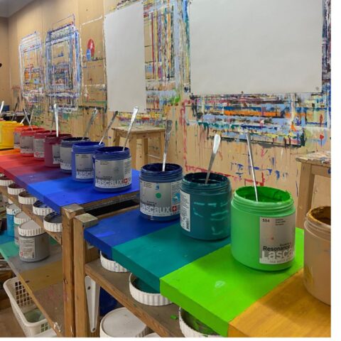 Atelierraum mit einer langen Holzbank, auf der geöffnete Farbtöpfe in Regenbogenreihenfolge von Gelb über Rot, Blau und Grün bis Beige stehen. In jedem Topf ste