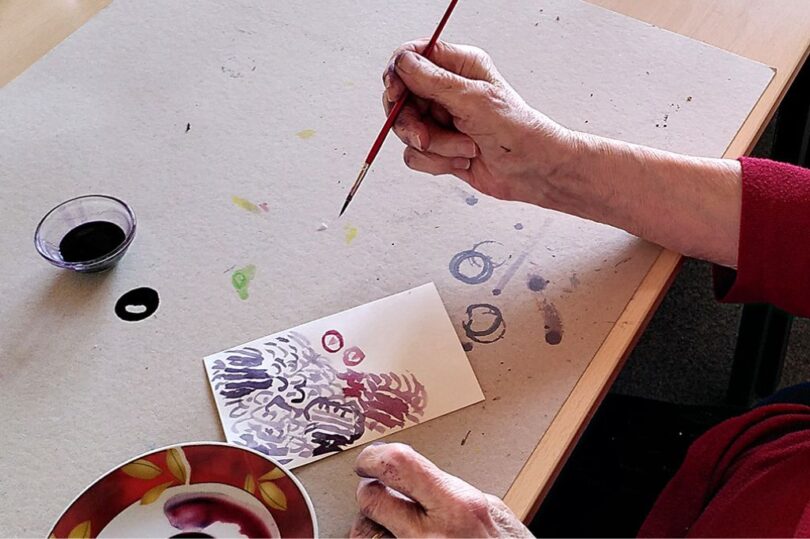Eine Person mit älteren Händen sitzt an einem Tisch und malt mit einem feinen Pinsel ein abstraktes Aquarellbild auf eine kleine Karte. Auf dem braunen Papierun