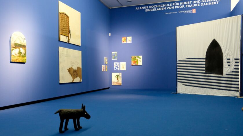Innenraum einer Kunstgalerie mit vollständig blauen Wänden und blauem Boden. Links hängen mehrere gerahmte Bilder, darunter Tier- und Pflanzenmotive. An der Rüc