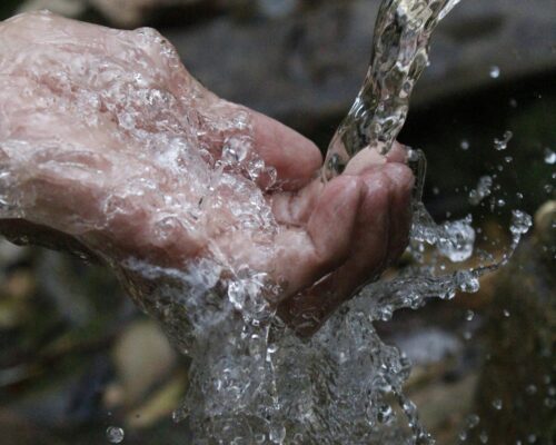Nahaufnahme einer menschlichen Hand, die klares, sprudelndes Wasser auffängt, während es darüber hinweg und zwischen den Fingern hindurchfließt.