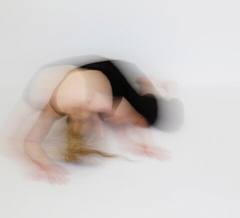 Verschwommene Aufnahme einer Person mit blondem Haar in schwarzer Sportkleidung, die sich auf einem hellen Boden nach vorn beugt, mit Stirn nahe dem Boden und a