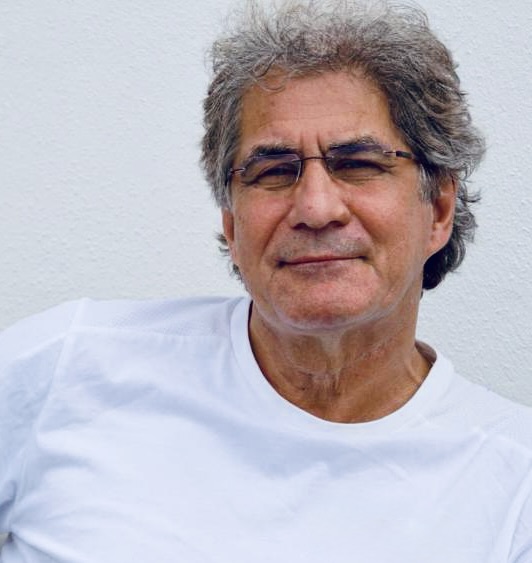 Älterer Mann mit grauen, lockigen Haaren und Brille lehnt entspannt vor einer weißen Wand und trägt ein schlichtes weißes T‑Shirt.