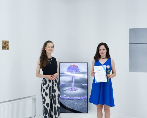 Preisverleihung in einer Kunstgalerie mit digitalem Kunstwerk im Hintergrund Zwei Frauen stehen in einem weißen Galerieraum nebeneinander. Links trägt die Frau ein schwarzes Top und eine weite Hose mit schwarz‑weißen Flecken, rechts träg