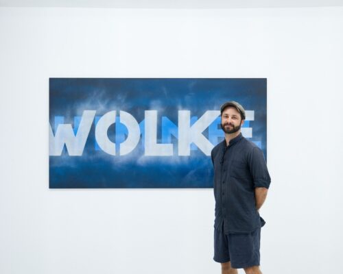 Mann posiert vor Kunstwerk mit dem Schriftzug „WOLKE“ in Galerie Ein Mann mit Bart steht in einem weißen Galerieraum vor einem großen rechteckigen Bild, auf dem in weißen Buchstaben das Wort „WOLKE“ auf blauem, wolkigem Hinte