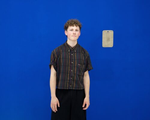 Person in weiter schwarzer Hose vor leuchtend blauer Wand Person mit lockigen kurzen Haaren steht vor einer kräftig blauen Wand. Sie trägt ein kurzärmliges, gestreiftes Hemd, sehr weite schwarze Hosen und Sandalen. Rec