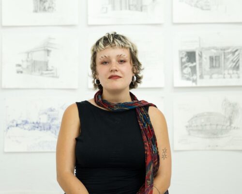 Porträt einer Person in einer Zeichen-Ausstellung Junge erwachsene Person mit kurzem, blondem Haar, schwarzem ärmellosen Oberteil, rotem Rock und buntem Schal steht in einer Galerie vor einer Wand mit mehreren