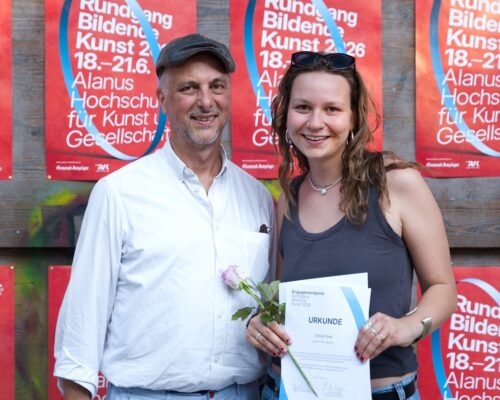 Auszeichnung mit Urkunde an der Alanus Hochschule Zwei lächelnde Personen stehen nebeneinander vor einer Holzwand mit roten Plakaten der Alanus Hochschule. Die Person rechts hält eine Urkunde und eine einzelne