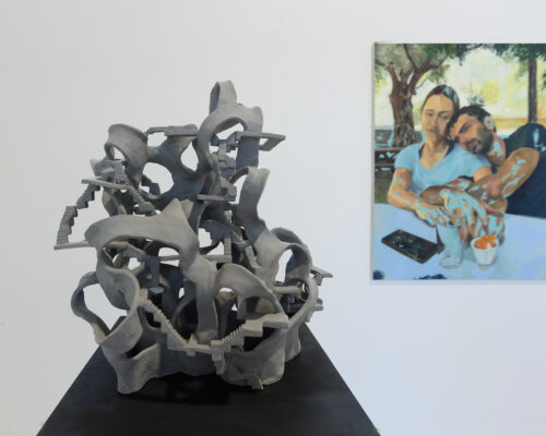 Galerieansicht mit einer grauen, abstrakten Skulptur im Vordergrund, die aus organisch wirkenden Formen und vielen kleinen Treppen besteht. Im Hintergrund hängt