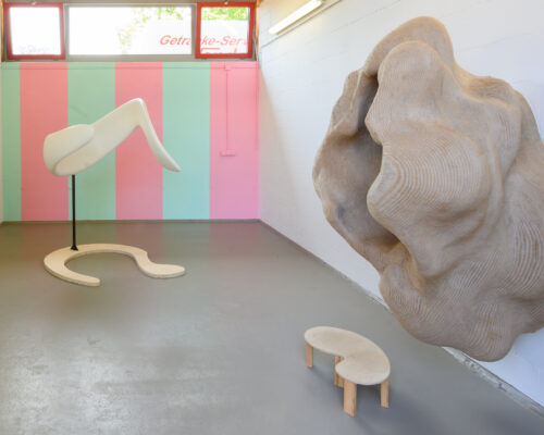 Innenraum einer Galerie mit pastellfarben gestreifter Wand in Rosa und Mintgrün. Links steht eine weiße, abstrakte Skulptur auf einem geschwungenen Sockel, die