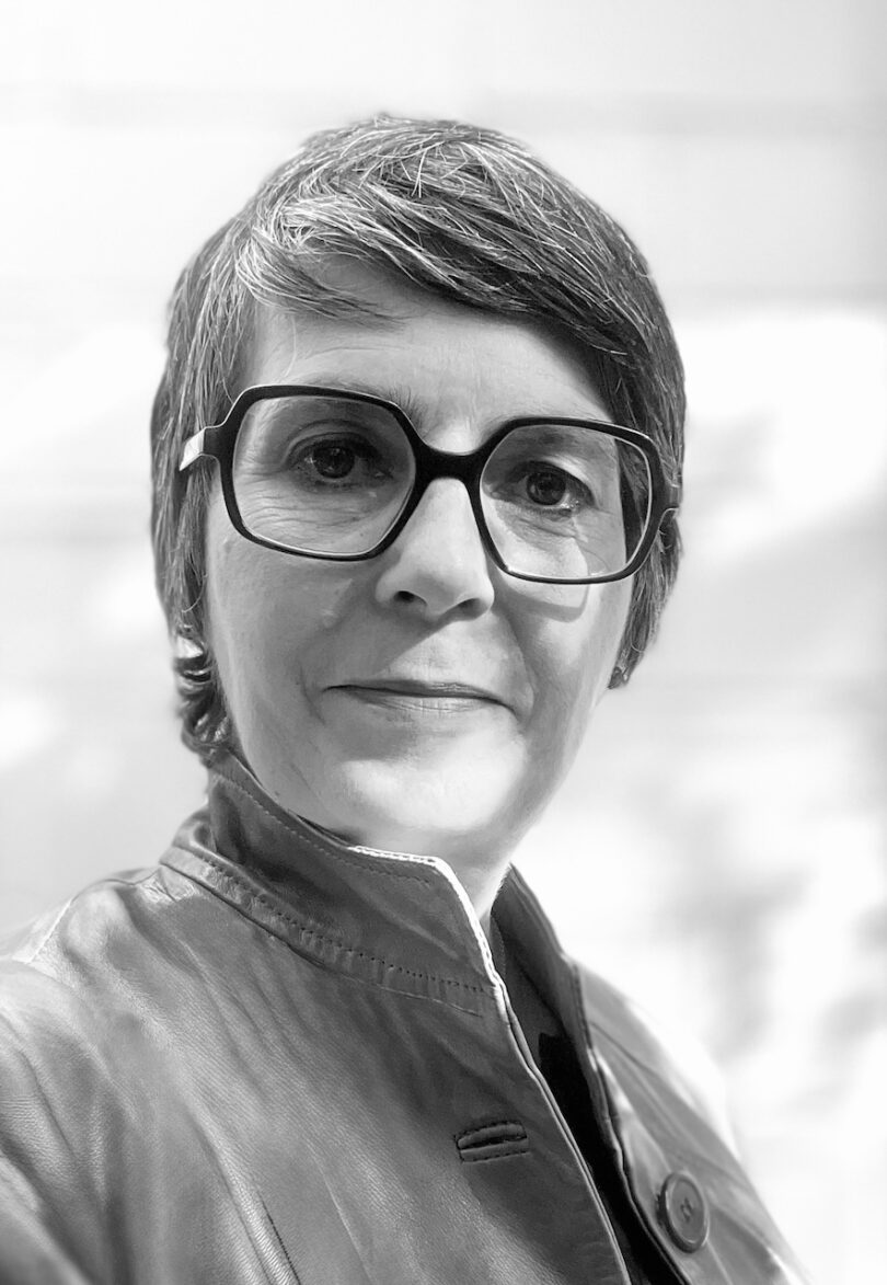 Schwarz-weiß-Porträt einer Person mit kurzem, seitlich frisiertem Haar und großer eckiger Brille, die mit ernstem, ruhigem Gesichtsausdruck in die Kamera schaut